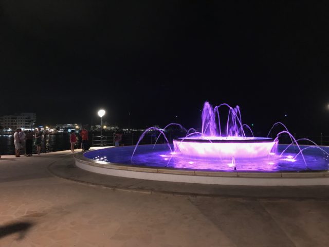 Purple Water Fountain By The Sea In The Night With People Standing And Watching The Water Jets