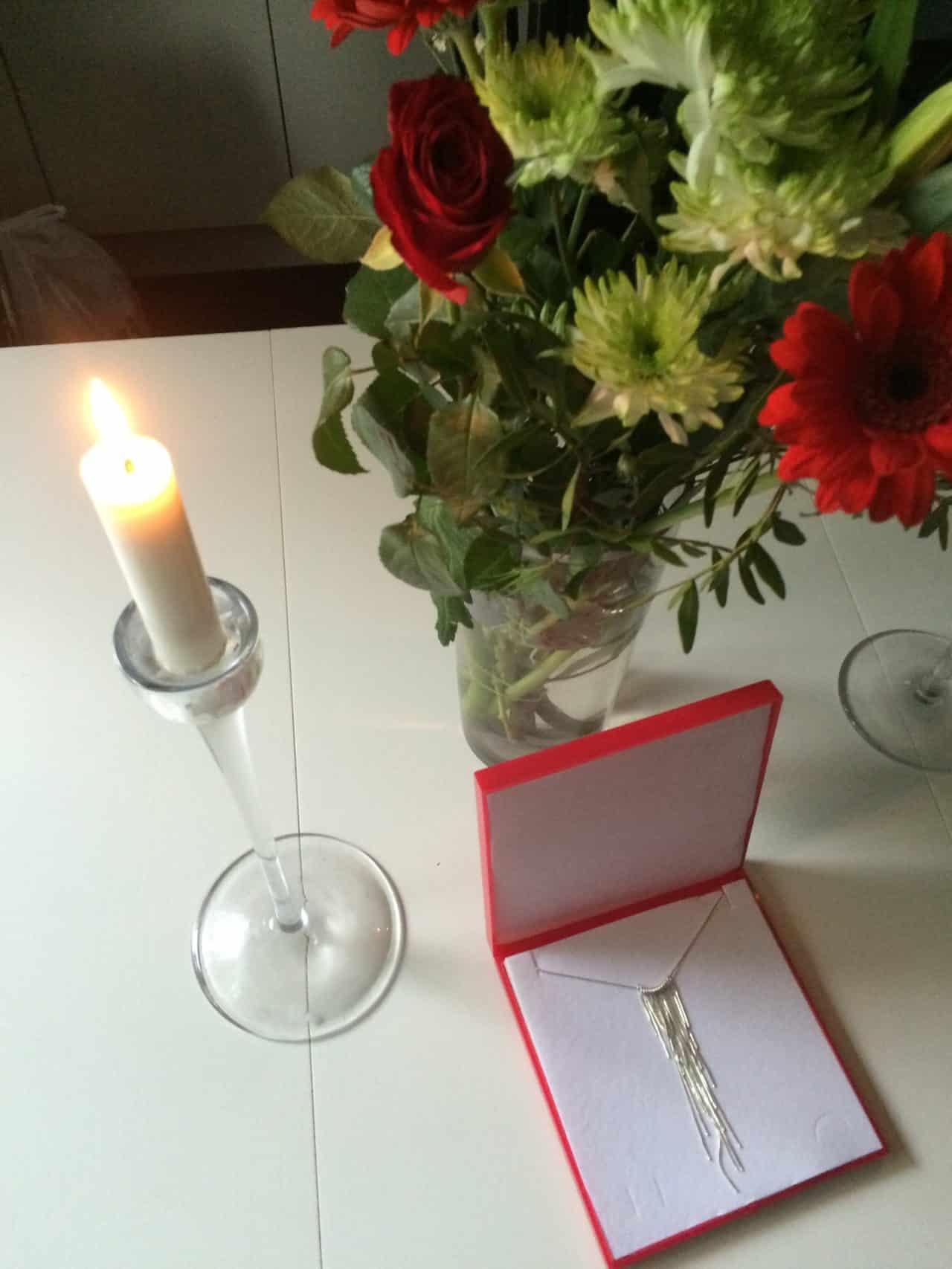 Silver Necklace In A Red Box On White Table With Flowers And Lighted Candle In Candlestick