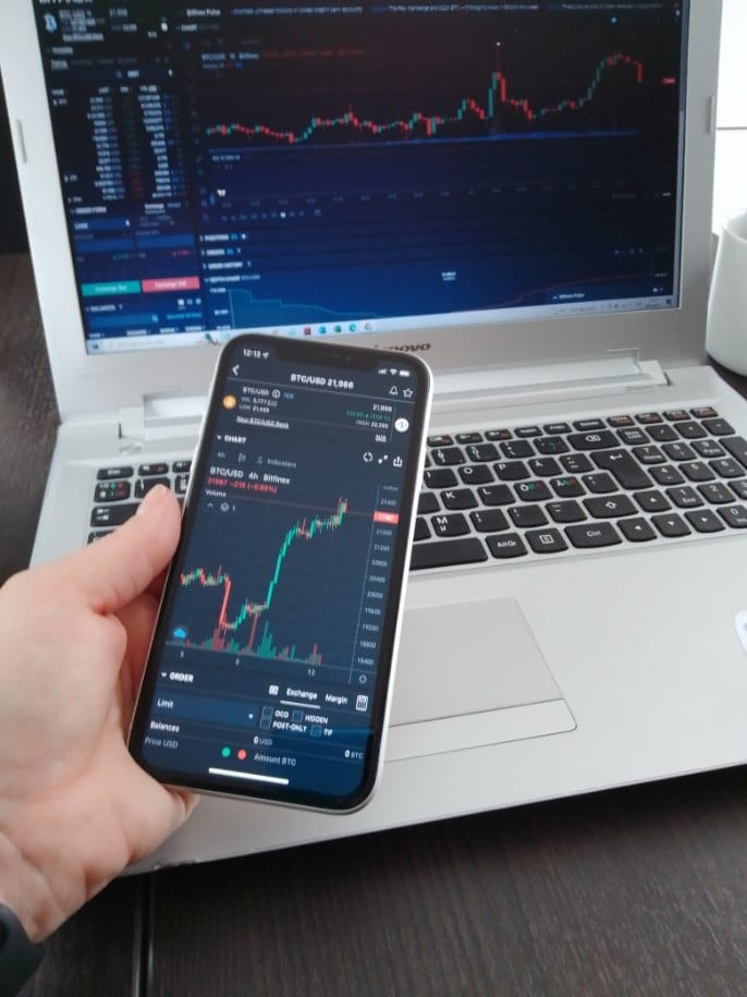 Hand Holding iPhone With Bitcoin Chart And Laptop With BTC
